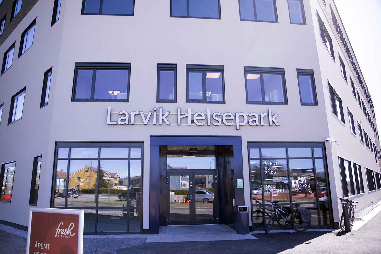 Inngangspartiet til Larvik Helsepark med skilt over døren. Bygget har lyse fasader og store vinduer. Tekst på vinduene viser åpningstider «05:00–24:00», «God trening» og «Fresh Fitness NO». En sykkel står parkert ved siden av inngangen, og nærliggende hus reflekteres i glasset. Bildet er tatt i klart dagslys.