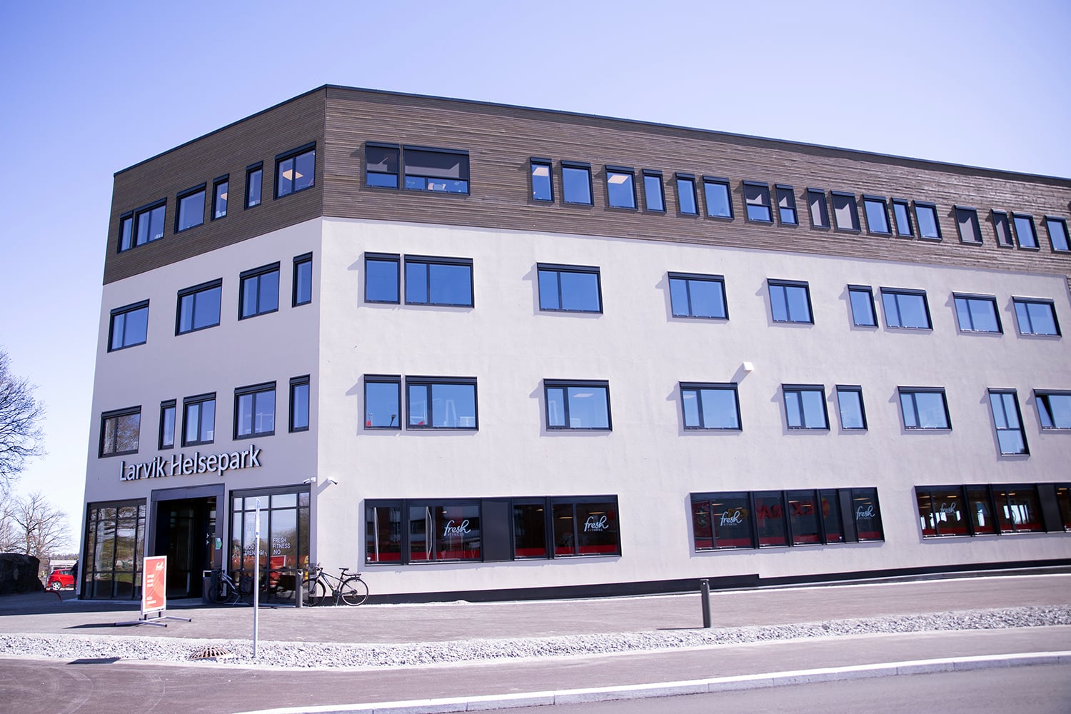 Stort, moderne bygg merket «Larvik Helsepark», sett fra siden og inngangspartiet. Fasaden er lys med en øvre etasje i tre, og mange jevnt plasserte vinduer. Ved inngangen står en sykkel, og gjennom vinduene er treningssenteret «Fresh Fitness» synlig. Asfaltert område og vei i forgrunnen, klart dagslys.