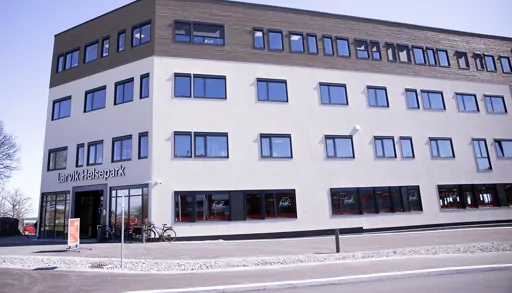 Stort, moderne bygg merket «Larvik Helsepark», sett fra siden og inngangspartiet. Fasaden er lys med en øvre etasje i tre, og mange jevnt plasserte vinduer. Ved inngangen står en sykkel, og gjennom vinduene er treningssenteret «Fresh Fitness» synlig. Asfaltert område og vei i forgrunnen, klart dagslys.