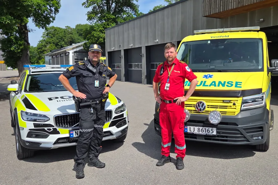 To menn i henholdsvis politiuniform og ambulanseuniform står foran politibil og ambulanse