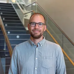 Person med briller står foran en innendørs trapp med glassrekkverk. Personen har på seg en lyseblå skjorte og er fotografert fra livet og opp. Trappen strekker seg oppover i bakgrunnen.