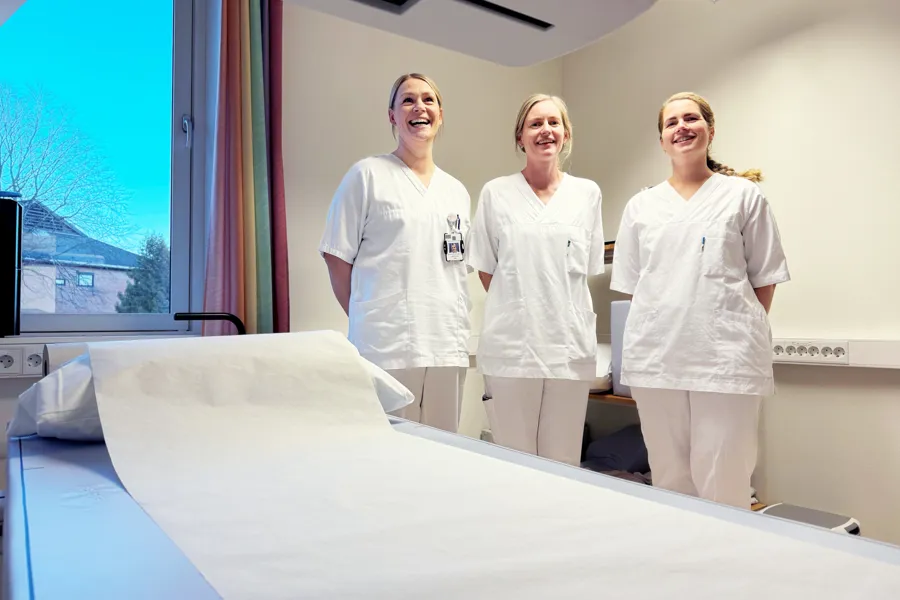 Tre kvinner i hvitt arbeidsantrekk står ved en pasientseng