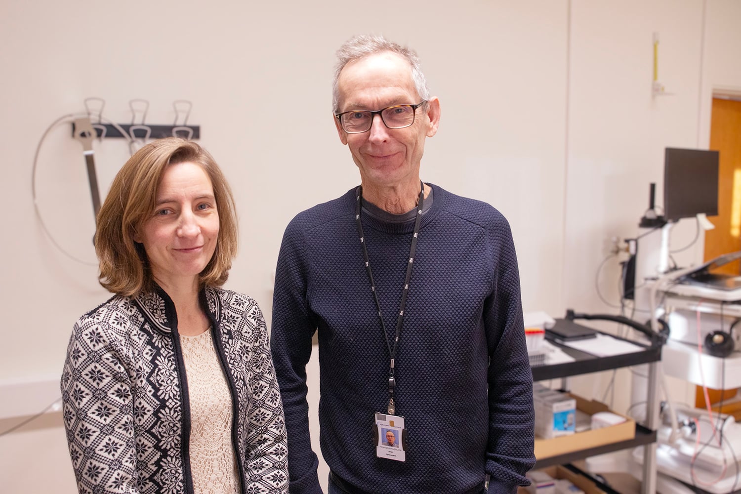 En mann og en kvinne står i et laboratorie- eller forskningsmiljø. De smiler mot kameraet, med teknisk utstyr, dataskjermer og hyller i bakgrunnen.
