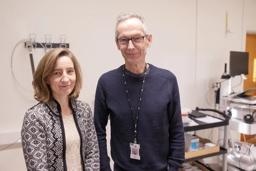 En mann og en kvinne står i et laboratorie- eller forskningsmiljø. De smiler mot kameraet, med teknisk utstyr, dataskjermer og hyller i bakgrunnen.