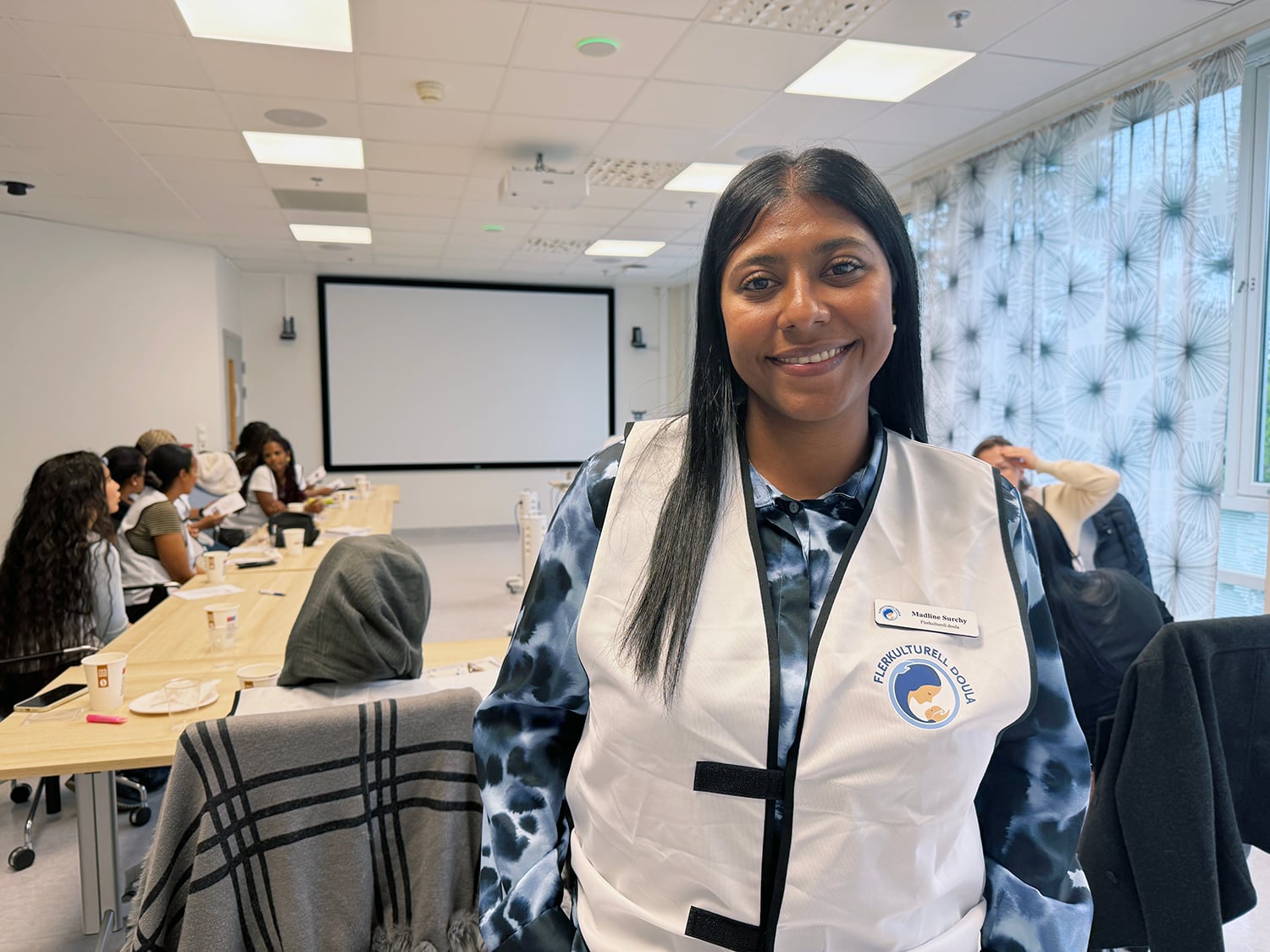 En kvinne står i forgrunnen av et møterom og har på en hvit vest med logoen «Flerkulturell Doula» og navneskiltet «Madline Sorchy». I bakgrunnen sitter flere deltakere rundt bord i U‑form, med kopper, papirer og penner foran seg. Rommet har lerret, taklys og mønstrede gardiner, og situasjonen viser en pågående opplærings- eller kursøkt.