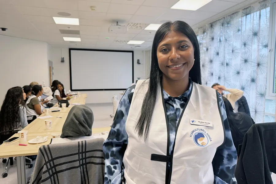 En kvinne står i forgrunnen av et møterom og har på en hvit vest med logoen «Flerkulturell Doula» og navneskiltet «Madline Sorchy». I bakgrunnen sitter flere deltakere rundt bord i U‑form, med kopper, papirer og penner foran seg. Rommet har lerret, taklys og mønstrede gardiner, og situasjonen viser en pågående opplærings- eller kursøkt.