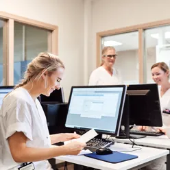Tre kvinner i hvitt arbeidstøy på et arbeidsrom med PCer