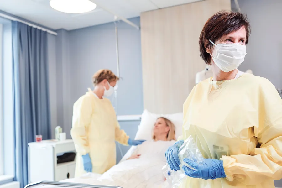 To sykepleiere med smittevernsutstyr står ved pasient som ligger i seng