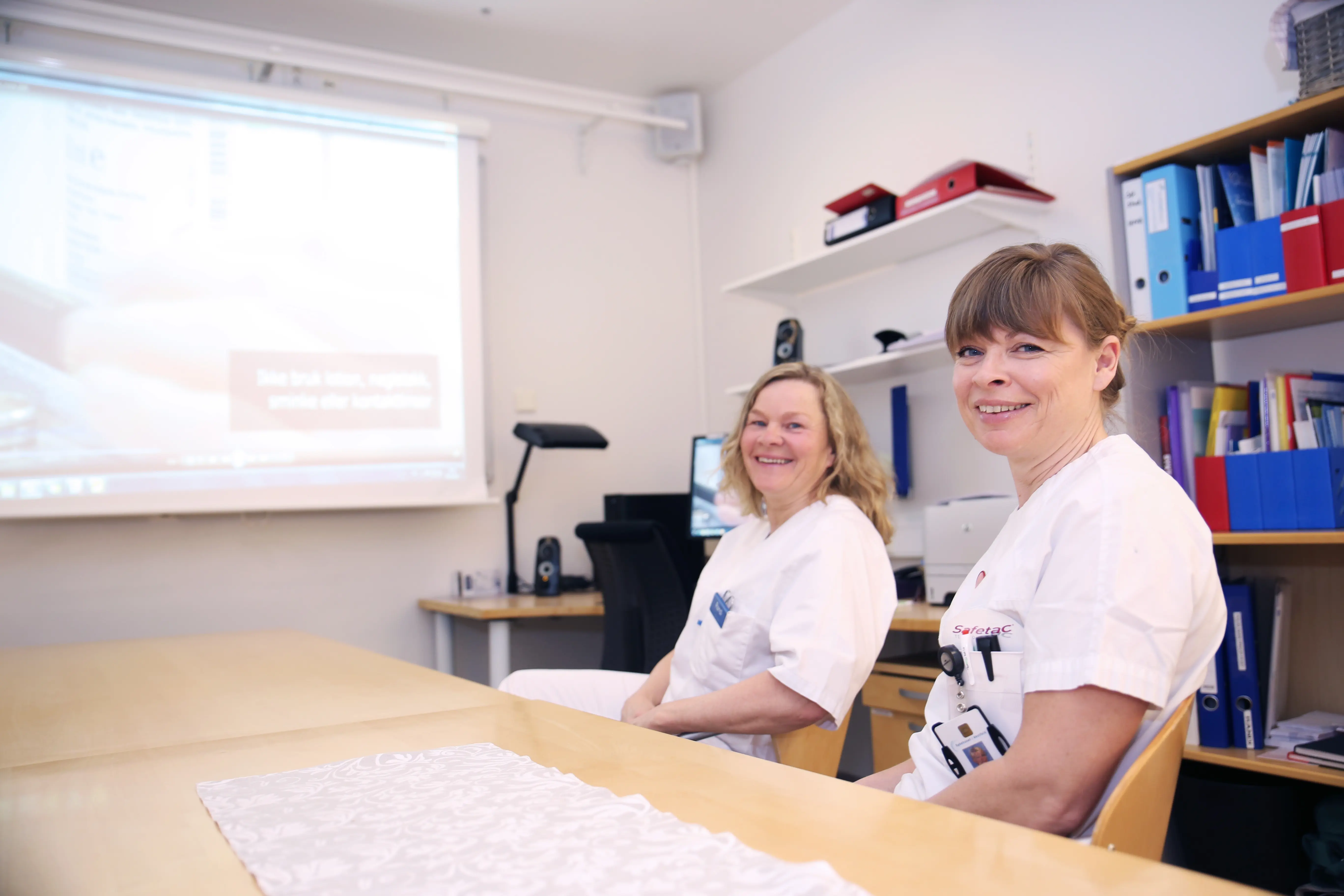 Sykepleierne Randi Oshaug og Pernille Simonsen sittende ved bordet med lerret i bakgrunnen hvor filmen går