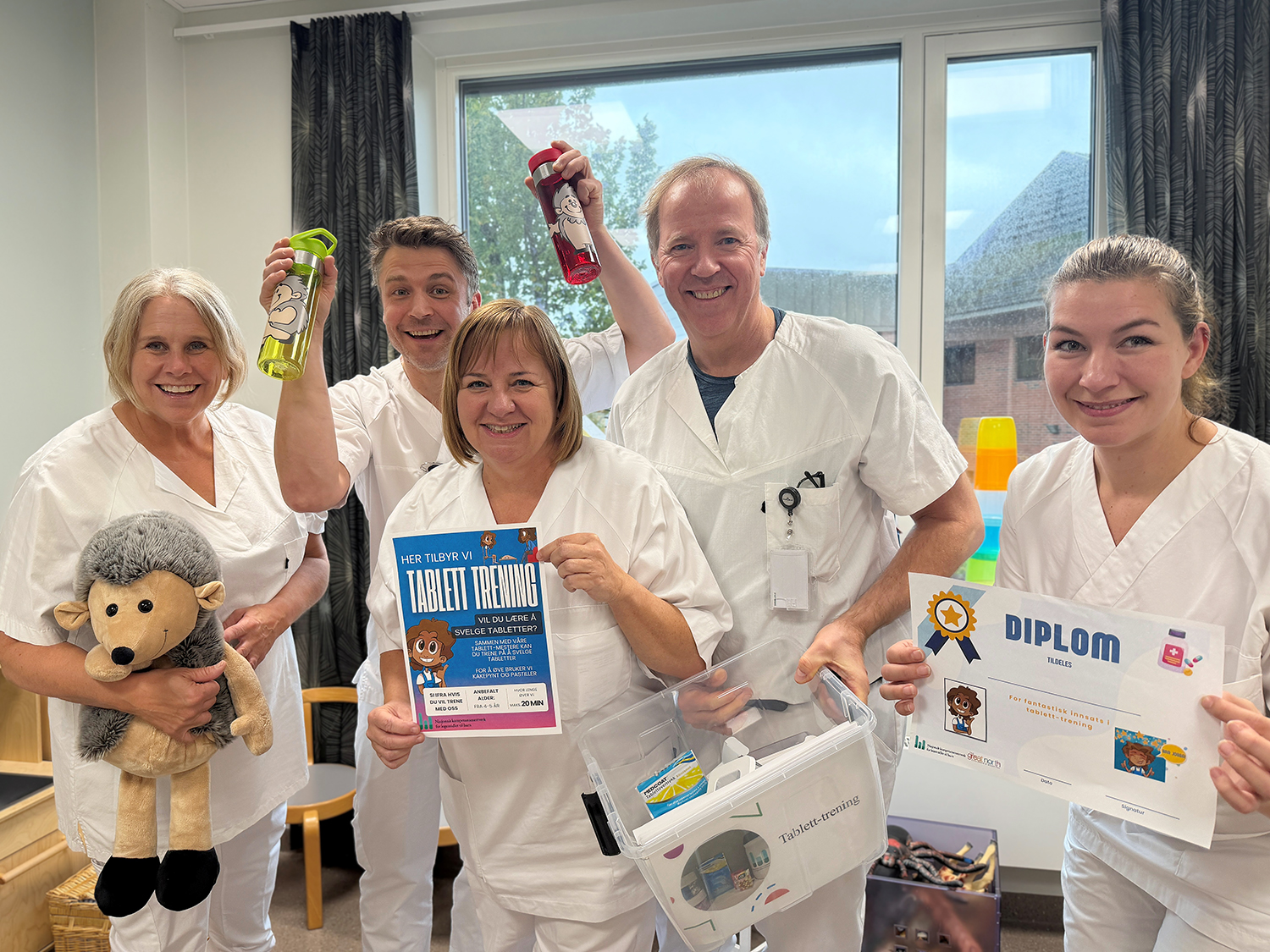 Fem ansatte i helsetjenesten står samlet i et rom og presenterer et opplegg for tablett-trening. I forgrunnen vises en informasjonsplakat med teksten «Her tilbyr vi tablett-trening», et diplom for gjennomført trening og en plastboks med opplæringsmateriell. To personer løfter fargerike drikkeflasker, og en person holder et kosedyr. Bildet illustrerer et engasjement for å hjelpe pasienter med å lære å svelge tabletter.
