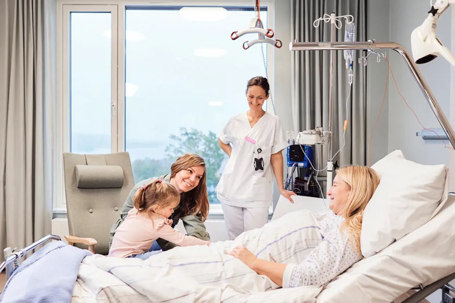 En pasient ligger i en seng og har besøk av en ung kvinne med et barn og en sykepleier